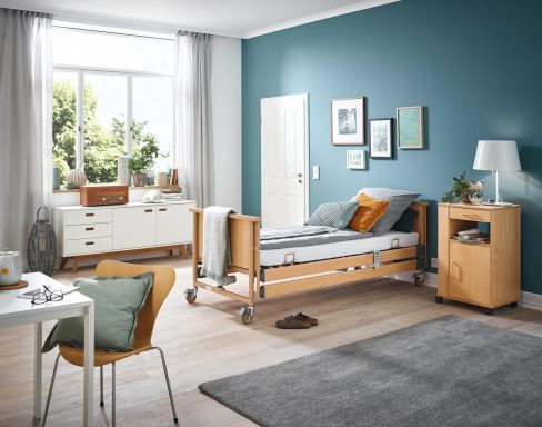 Helles Schlafzimmer mit Pflegebett, Holzmobiliar und bläulicher Wandfarbe.
