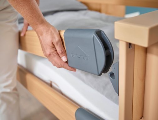 Eine Hand hebt einen grauen Bettseitenschutz an einem Holzbett an.
