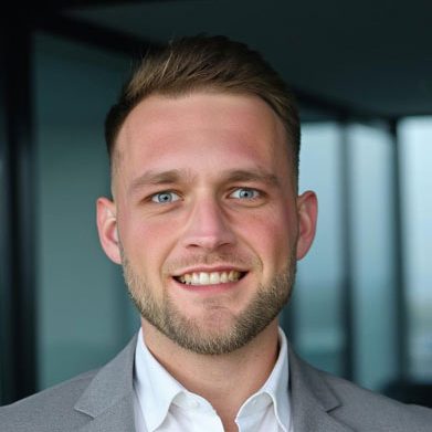 Lächelnder Mann mit kurzem, blondem Haar in einem grauen Anzug vor einer modernen Kulisse.