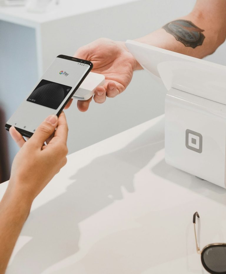 Hand, die ein Smartphone hält, übergibt ein Zahlungsmittel an eine Kasse.