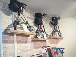 Drei rollstuhlarte Mobilitätshilfen auf Regalen an einer Ziegelwand.