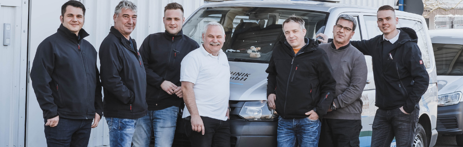 Sechs Männer stehen neben einem Fahrzeug, freundliche Ausdrücke und lässige Kleidung.
