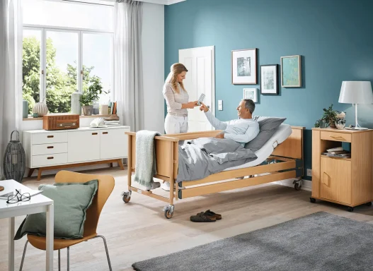 Pflegekraft hilft einem Patienten im Bett in einem modernen, hellen Zimmer.