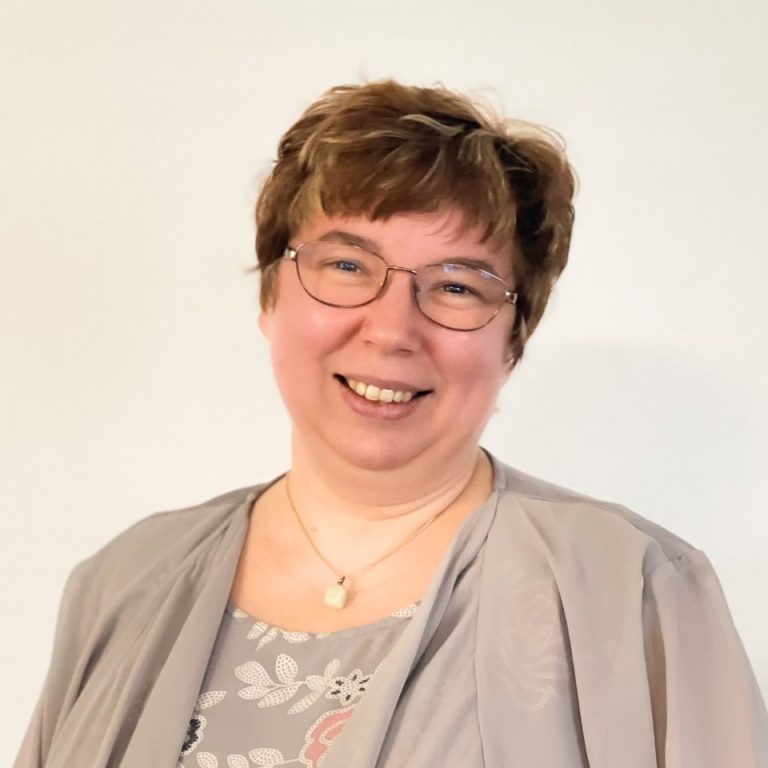 Lächelnde Frau mit Brille, kurzhaarig, in grauem Oberteil vor hellem Hintergrund.